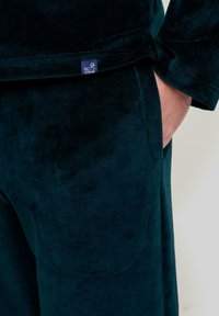 Top de terciopelo verde oscuro con mangas largas, que presenta un bolsillo y una pequeña etiqueta azul con un logo de pato en el dobladillo. Textura suave.