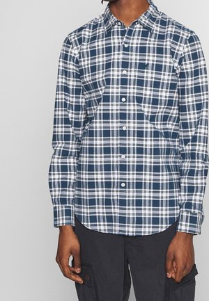 Homme portant une chemise à carreaux bleue, blanche et verte à boutons avec manches longues et un pantalon cargo sombre.