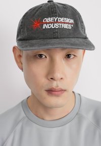 Gray cap with "OBEY DESIGN INDUSTRIES" embroidered in white and red, featuring a six-panel design. Soft fabric with a curved brim.