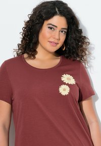 Camiseta de manga corta color marrón en tejido suave con dos flores bordadas en crema en el lado izquierdo. Cuello redondo y corte relajado.