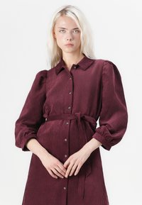 Burgunderfarbene Cordshirtkleid mit Kragen, Knopfverschluss, Taillengürtel und puffärmeln mit Raffung.