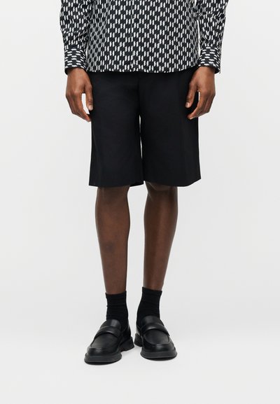 Personne portant un short noir jusqu'aux genoux, des chaussettes noires, des chaussures noires sans lacets, et une chemise à manches longues à motif noir et blanc.