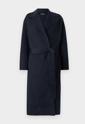 Cappotto lungo in lana blu navy con ampi revers e cintura in tessuto abbinato annodata in vita su sfondo bianco.