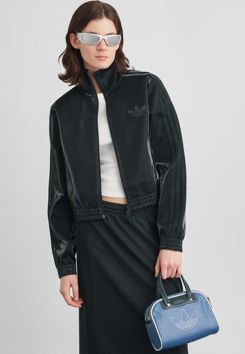 Casaco de vinil preto com cintura elástica, três riscas nas mangas, combinado com uma saia preta e uma mala azul com detalhes brancos.