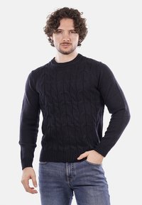 Uomo con capelli ricci che indossa un maglione scuro a trecce e jeans blu, in piedi con una mano in tasca davanti a uno sfondo bianco.