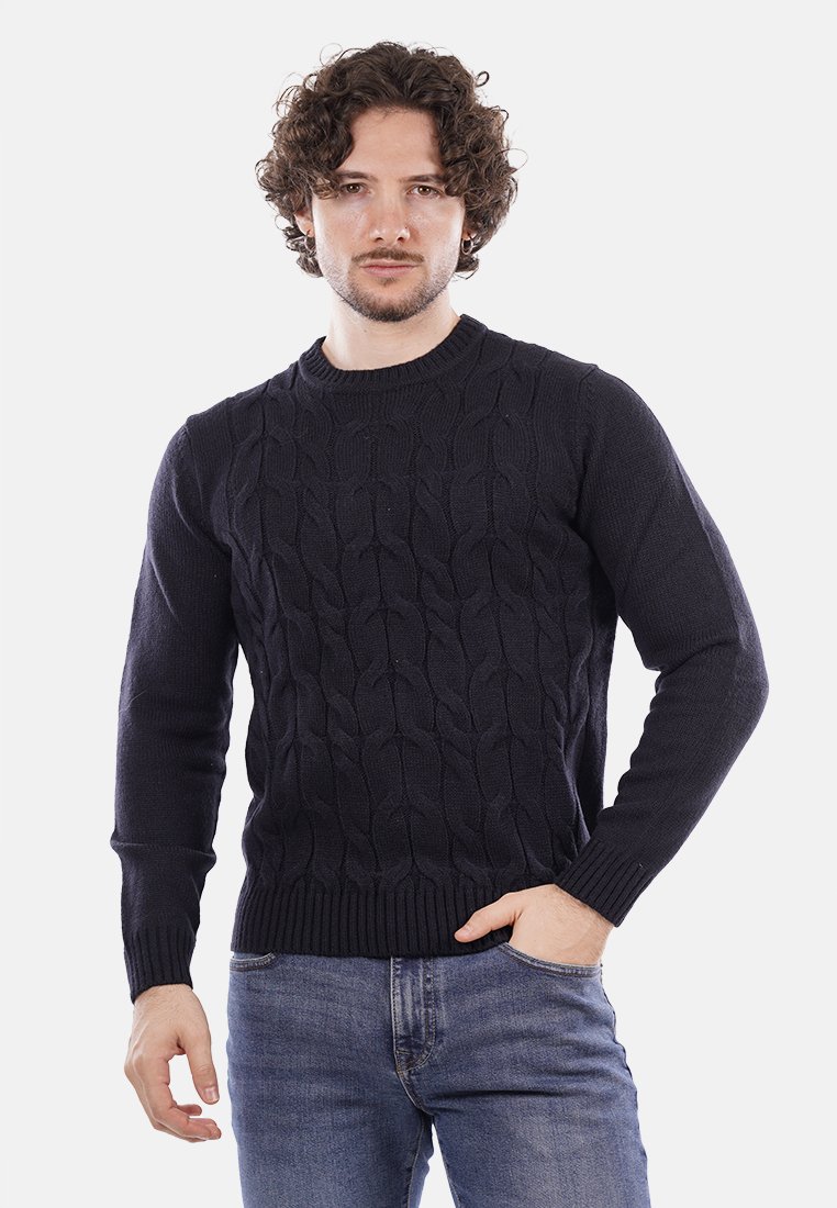 Uomo con capelli ricci che indossa un maglione scuro a trecce e jeans blu, in piedi con una mano in tasca davanti a uno sfondo bianco.