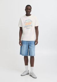 Kremkrāsas t-krekls ar zilu un apelsīnu grafiku, uz kuras rakstīts "DENIM AUTHENTIC IN STYLE", kombinēts ar džinsu šortiem un pelēkiem sporta apaviem.