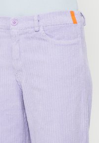 Pantalones de pana color púrpura claro con textura acanalada, cierre de botón y un detalle naranja en la cintura.