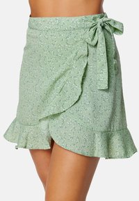 Zelená květinová zavinovací sukně vyrobená z lehkého materiálu, s volánkovým lemem, detailem mašle a malými bílými květinovými vzory.