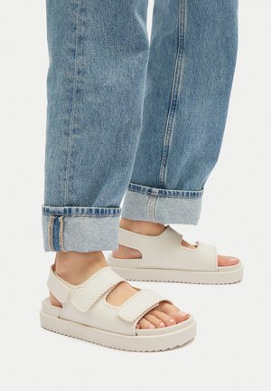 Jasnobeżowe sandały z regulowanymi paskami, masywną białą podeszwą i teksturowaną krawędzią, zestawione z niebieskimi jeansami podwiniętymi na dole.