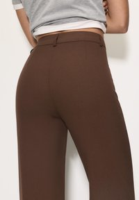 Bruine, hoog-waist broek met een gladde textuur, wijd uitlopende pijpen en minimale stiksel-details. De stof lijkt lichtgewicht en vloeiend.