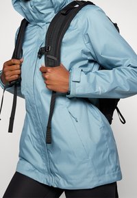 Vaaleansininen vedenpitävä takki hupulla, jossa on säädettävät hihansuut, vetoketjulliset taskut, ja se on puettu mustien reppihihnojen päälle.