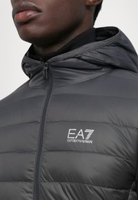 Veste grise matelassée avec capuche, fermeture éclair et coutures verticales. Équipée d'un logo EA7 brodé sur la poitrine gauche. Texture lisse.