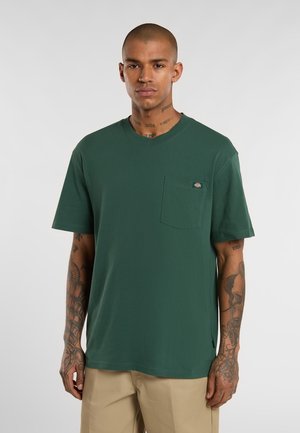 Dunkelgrünes Baumwoll-T-Shirt mit Rundhalsausschnitt, kurzen Ärmeln und einer Brusttasche auf der linken Seite. Verfügt über ein kleines Logo-Tag an der Tasche. Glatte Textur.