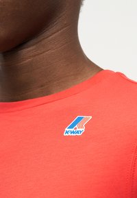 Κοντινό πλάνο του λαιμού και του ώμου ενός ατόμου που φοράει φωτεινό κοραλλί πουκάμισο με το λογότυπο K-Way στο πάνω μέρος του στήθους.