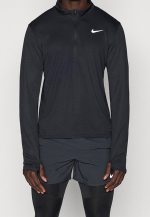 Homme portant un haut Nike noir à manches longues avec fermeture éclair et logo blanc, short noir et leggings noirs, debout les bras relâchés le long du corps.
