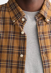 Koszula w kratę z guzikami, posiadająca brązowy, musztardowy i niebieski wzór, z zaokrąglonym kołnierzykiem i białymi guzikami, nałożona na szary T-shirt.