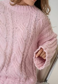 Gaiši rozā adīta džemperis ar augstu apkakli un pārmērīgiem piedurknēm, kuram ir teksturēti raksti un pūkains apdare.