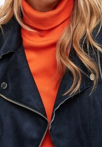 Giacca in suede navy con hardware argentato, indossata sopra un dolcevita a coste arancione. Tessuti testurizzati con dettagli di cucitura visibili.