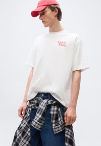 Άσπρο βαμβακερό μπλουζάκι με κόκκινο λογότυπο "Calvin Klein", συνδυασμένο με μπλε τζιν και καρό φανέλα σε ασπρόμαυρο που έχει δεθεί στη μέση. Κόκκινο καπέλο.