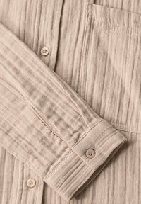 Světle béžová texturovaná košile s pomačkaným vzhledem, s knoflíkovým manžetou a kapsou na prsou. Jednoduchý, funkční design.