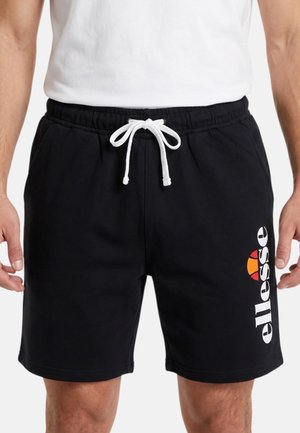 Homme portant un short noir avec cordon blanc et logo "ellesse" sur la jambe gauche, associé à un t-shirt blanc uni, montré de la taille vers le bas.
