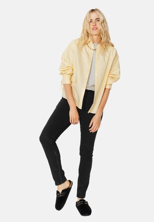 Chemise rayée jaune à boutons portée sur un haut gris, jean skinny noir et chaussons noirs avec des accents décoratifs. Tenue décontractée.