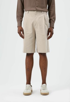 Beige op maat gemaakte shorts met een ontspannen pasvorm, voorplooien en zijzakken, gecombineerd met witte sneakers met beige accenten.