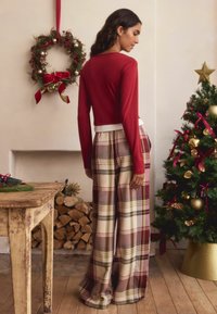Maroon long-sleeve top paired with wide-leg plaid pants in cream, red, and green. Wooden table with logs and a decorated Christmas tree nearby.