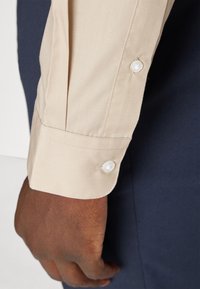 Beige Hemdmanchette mit Knopfakzenten, glatter Textur und maßgeschneidertem Schnitt. Die Hand zeigt einen dunklen Stoff im Hintergrund.