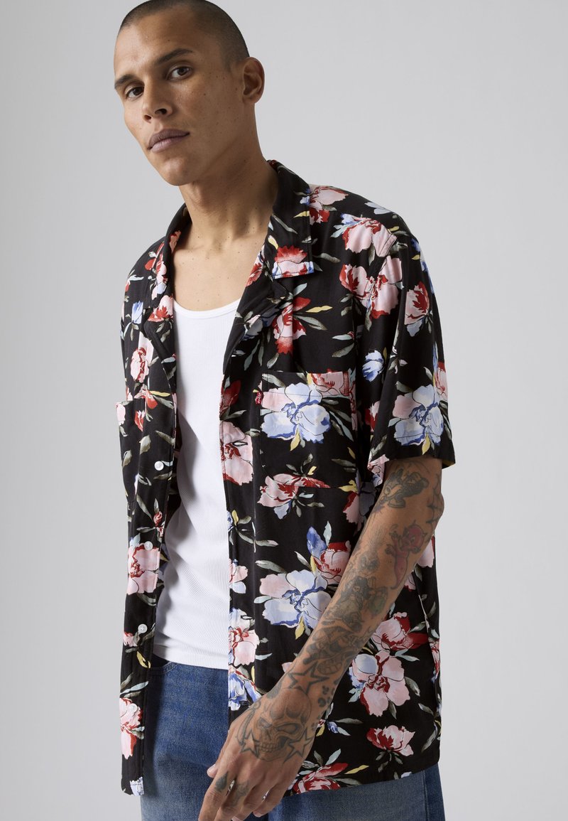 Ung mand med kort hår bærer en sort blomster skjorte over en hvid tanktop og viser sin tatoverede højre underarm mod en ensartet lys baggrund.