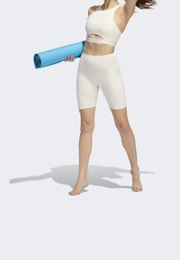 Bílý sportovní set s krátkým topem a šortkami s vysokým pasem. Modelka drží modrou jógovou podložku, která ukazuje hladký, těsný design.