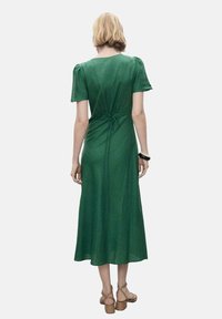 Rochie verde, de lungime midi, cu mâneci scurte tip puff, fundă la talie și o textură netedă. Poartă sandale bej cu bretele.