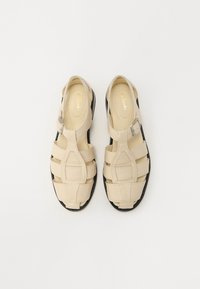 Sandale bej cu un design deschis, având curele late și o cataramă. Talpa neagră oferă un contrast. Textură netedă, stil casual.