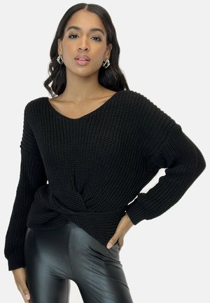 Maglione nero a maglia con scollo a V, vestibilità ampia e dettaglio frontale attorcigliato, abbinato a leggings in ecopelle neri, che mettono in mostra texture e design.