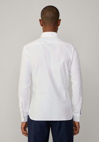 Camisa blanca de manga larga con cuello estándar, puños abotonados y una textura suave. La vista trasera muestra un corte recto y un pequeño detalle en forma de lazo.