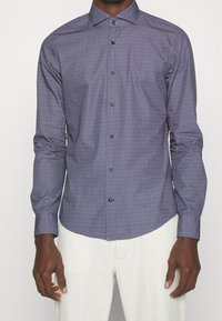 Man in een getailleerde overhemd met lange mouwen, voorzien van een subtiel geometrisch patroon, en witte broeken, staande met ontspannen armen.