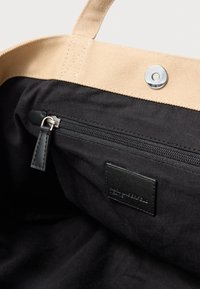 Drobės rankinė su šviesiai ruda išore ir juoda vidine dalimi. Turi užtraukiamą kišenę ir metalinę spaudės užsegimą. Tekstūruota paviršius.