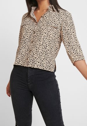 Femme portant une chemise beige à imprimé léopard avec boutons et manches trois quarts, et un jean skinny noir taille haute, sur fond uni.