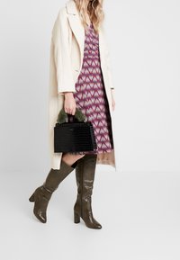 Bolso negro de cuero con relieve de cocodrilo y asa de piel de foca verde, combinado con un vestido estampado y botas de caña alta verde oliva, bajo un abrigo color crema.