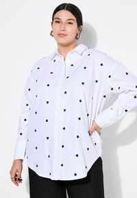 Witte button-up shirt met zwarte hartpatronen, met lange mouwen en een gebogen zoom. Gemaakt van lichtgewicht, stevig stof.