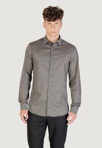Giovane uomo con capelli ricci che indossa una camicia a maniche lunghe grigia con motivo e pantaloni neri, in piedi su uno sfondo neutro.