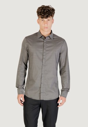 Joven con cabello rizado que lleva una camisa de botón de manga larga gris con patrón y pantalones negros, de pie contra un fondo liso.