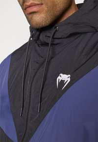 Veste coupe-vent noire et bleue avec capuche, fermeture éclair, tissu texturé et un accent de logo blanc sur la poitrine.