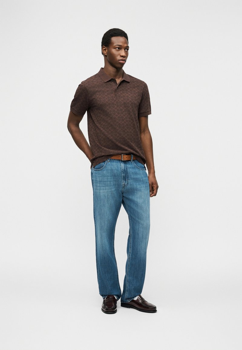 Man standing with hands in pockets, wearing a patterned brown polo shirt, blue jeans, brown belt, and dark brown shoes.