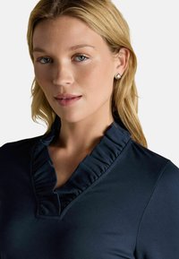 Mornarsko modra majica z nabrano ovratnikom, mehko teksturo in sproščenim krojem. Pogleduje bližnji posnetek, ki se osredotoča na dekolte in zgornji del tkanine.