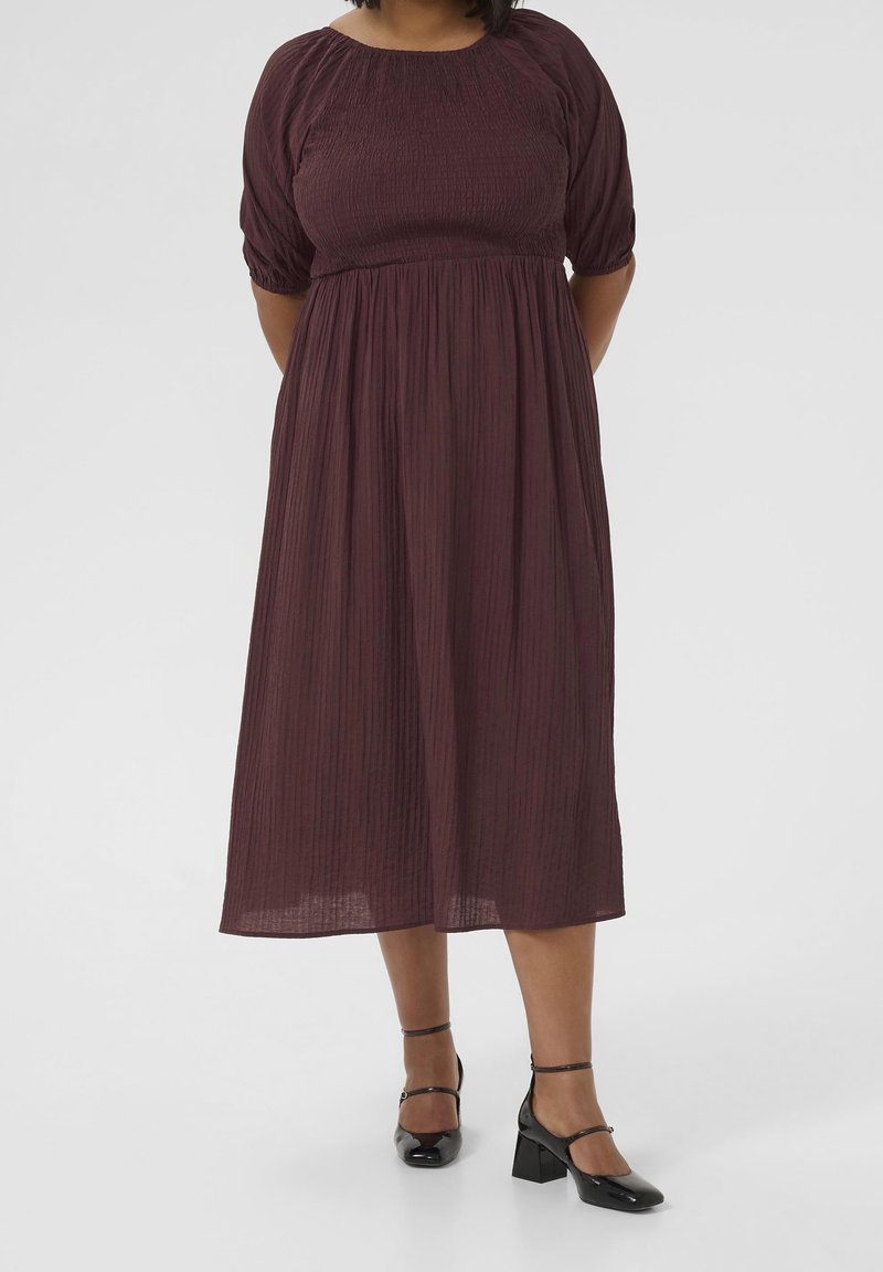 Femme portant une robe midi violet foncé avec des manches bouffantes et des chaussures à talons blocs noires avec bride à la cheville, debout devant un fond uni.