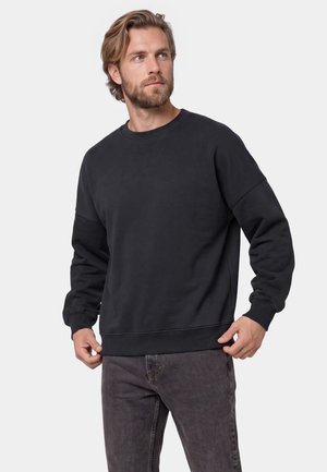 Mand med skæg og halvlangt hår iført en sort sweatshirt med rund hals og mørkegrå jeans, der kigger til siden.