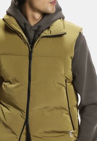 Mann, der eine olivgrüne Steppweste über einem braunen Hoodie trägt, nach rechts schaut, mit geschlossener Weste und sichtbaren Zugbändern am Hoodie.