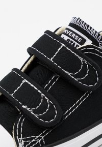 Sneaker nera in tela con cuciture bianche, due cinturini in velcro e una superficie testurizzata. Presenta un'etichetta con il logo sulla linguetta.
